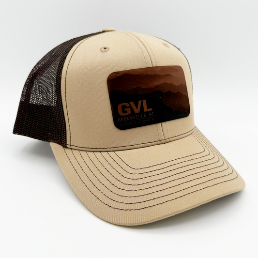 Beige and brown trucker cap with 'GVL' and Greenville, SC over a monochromatic mountain landscape view