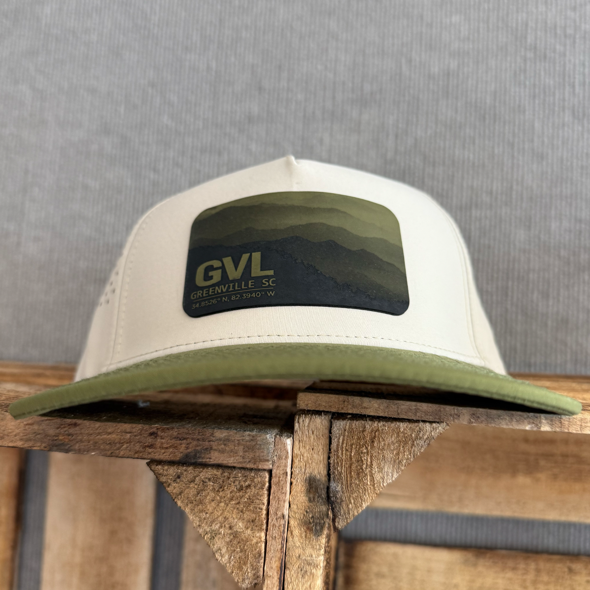 Beige and green trucker cap with 'GVL' and Greenville, SC over a monochromatic mountain landscape view