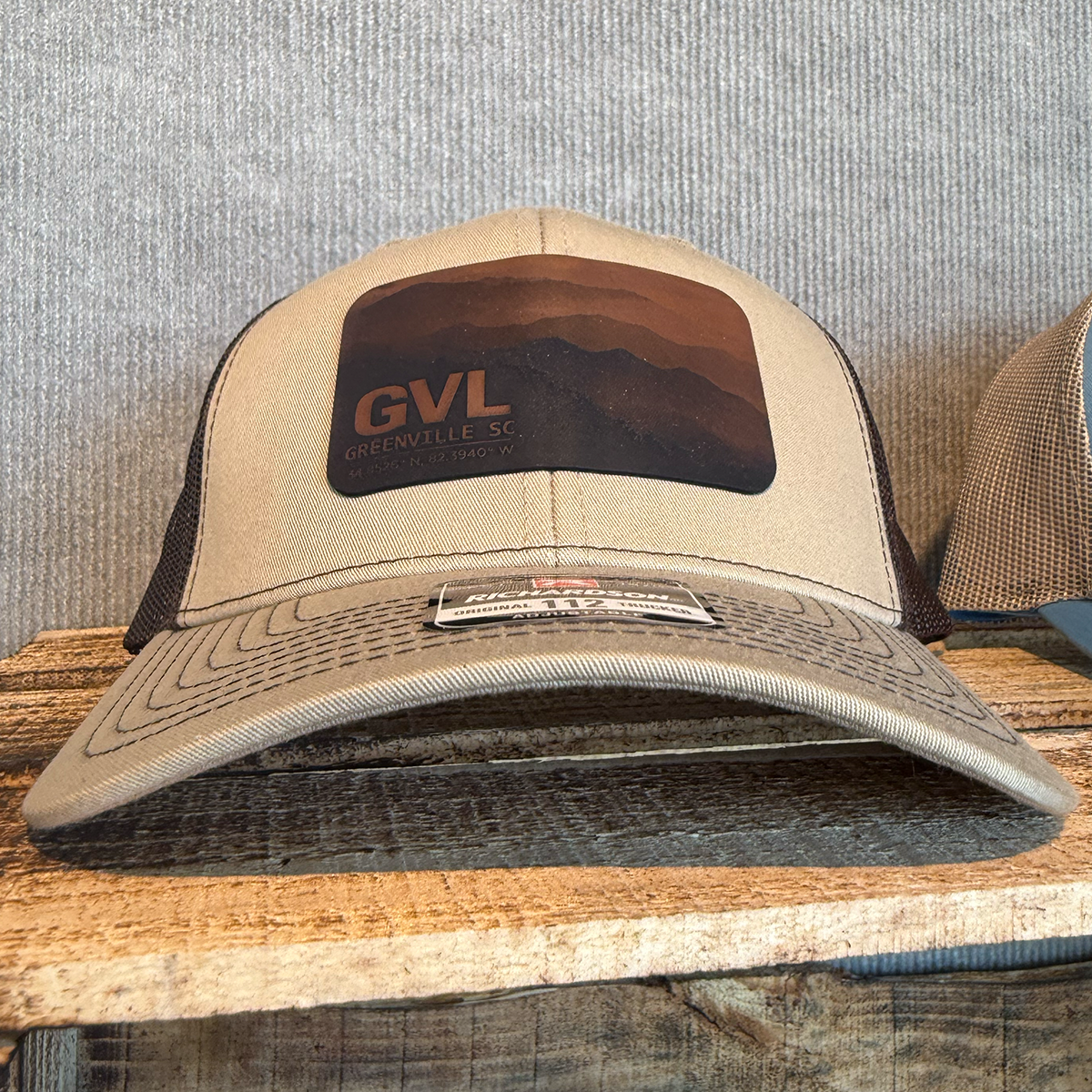 Beige and brown trucker cap with 'GVL' and Greenville, SC over a monochromatic mountain landscape view
