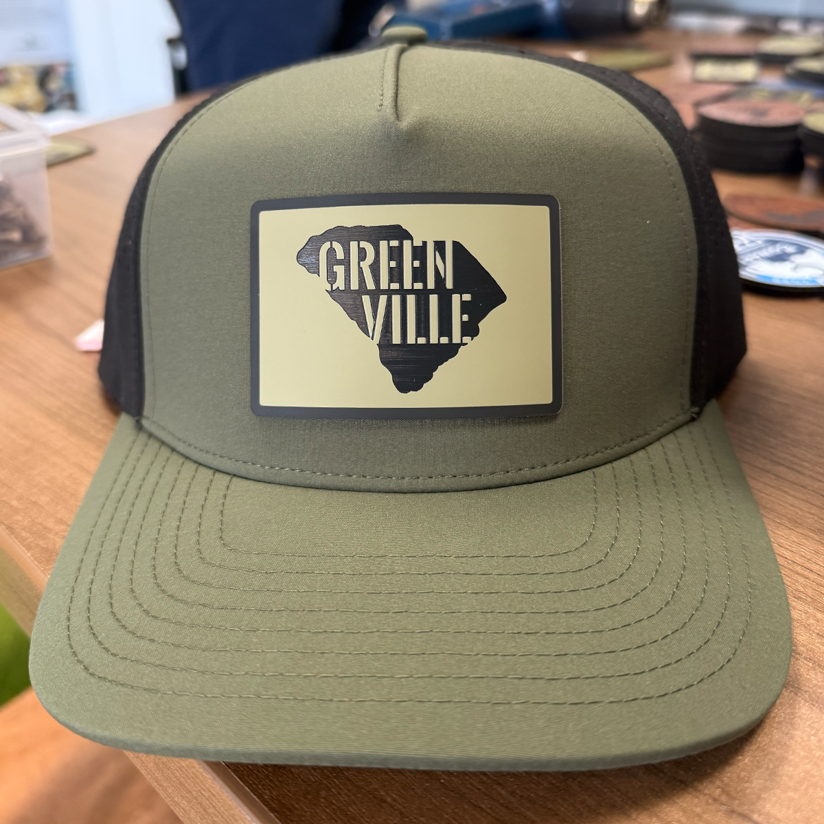 Green and black cap with 'Greenville' over a South Carolina state shape on wood surface.
