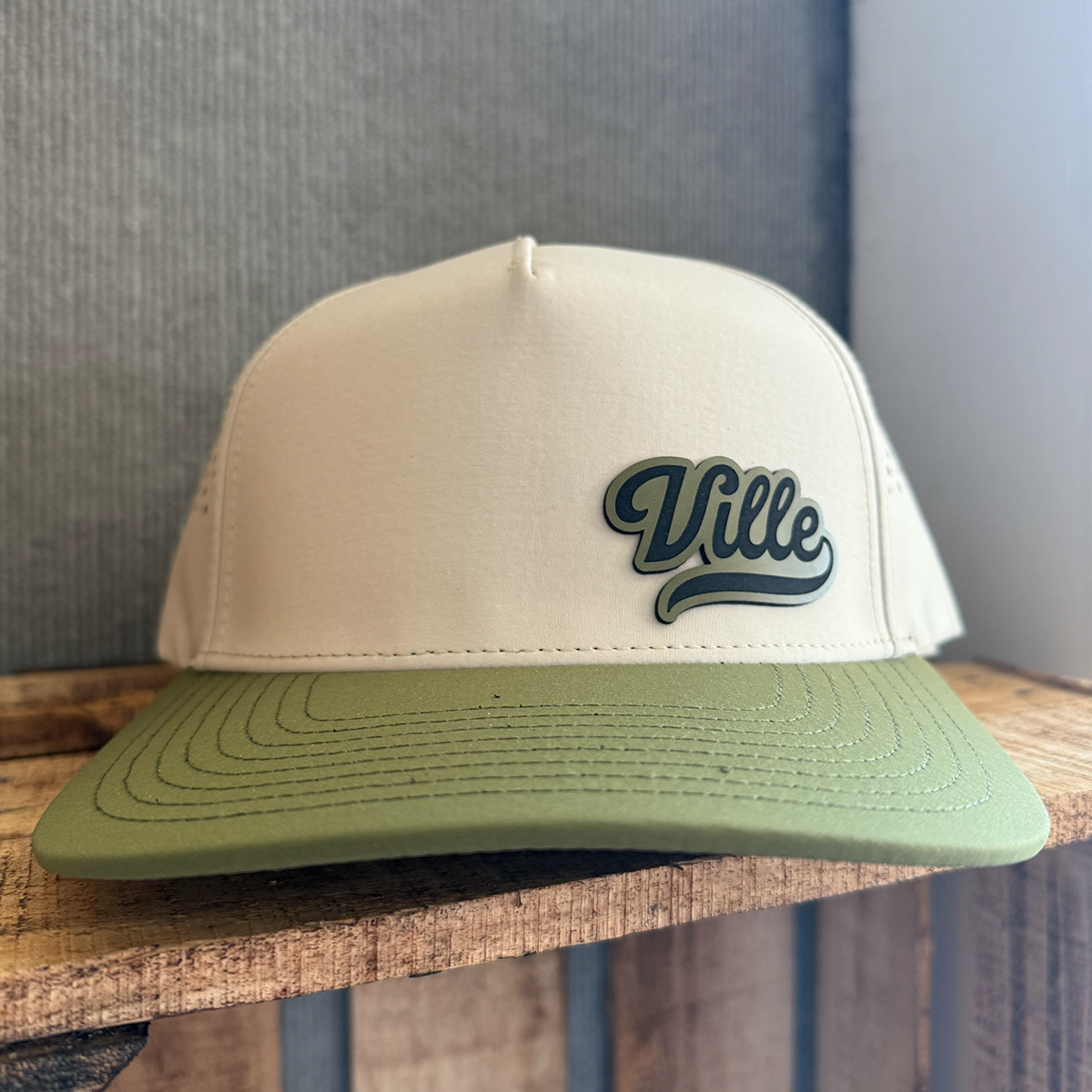 Beige and green cap with 'Ville' logo on a wooden surface