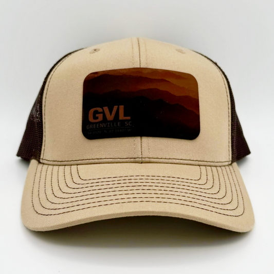 Beige and brown trucker cap with 'GVL' and Greenville, SC over a monochromatic mountain landscape view